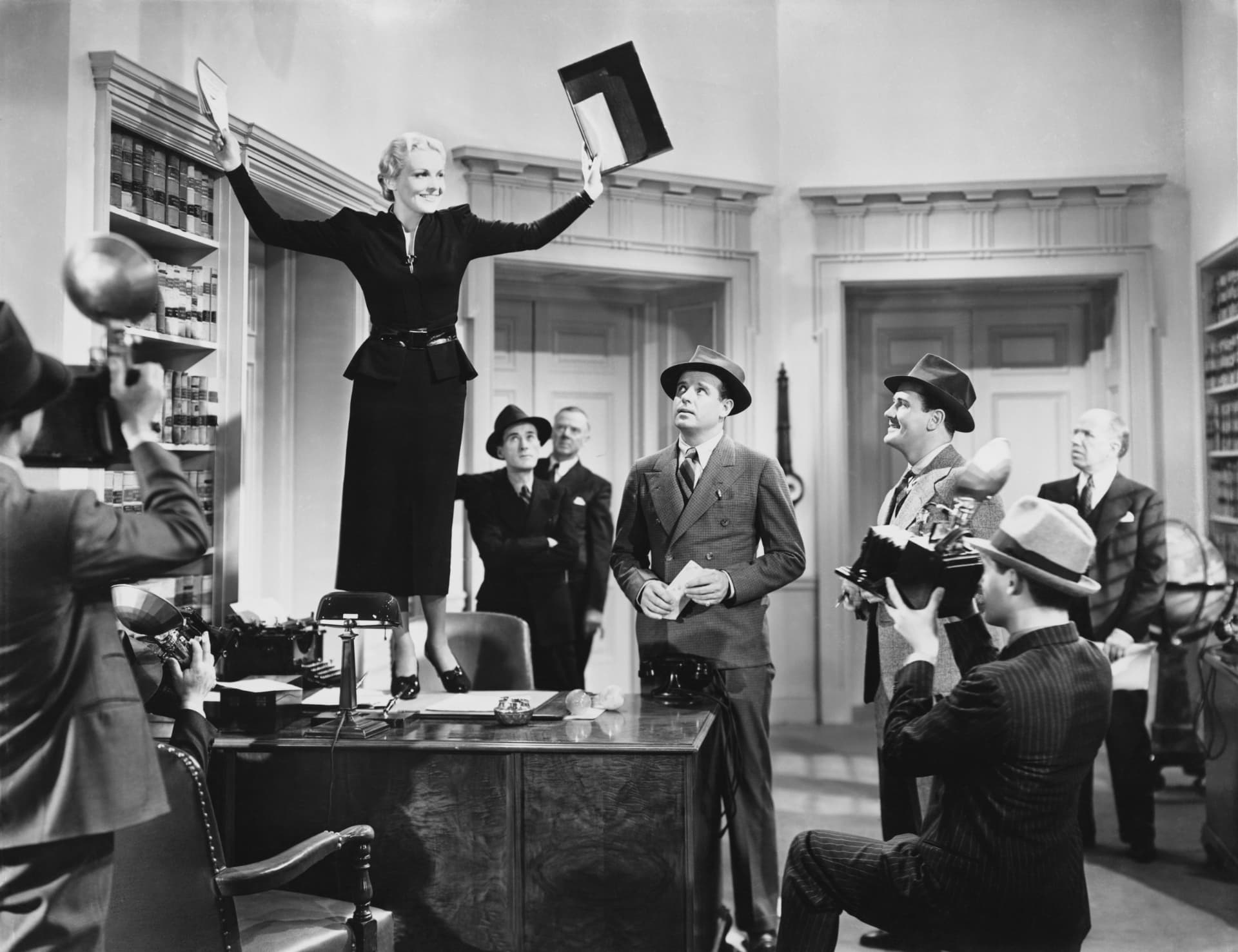 A woman commanding a busy office from atop a desk