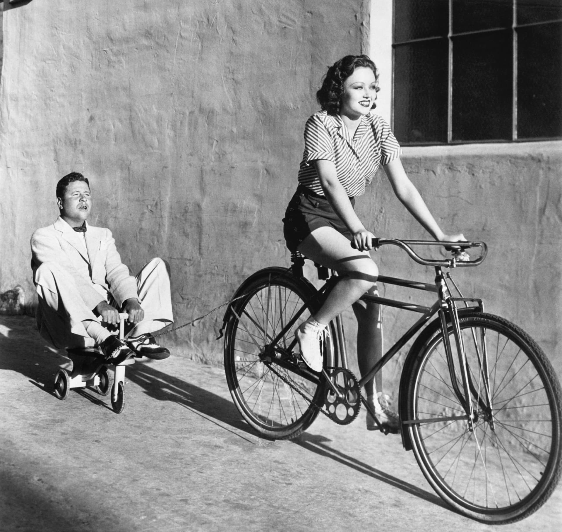 A woman on a bicycle towing a seated man on a small cart
